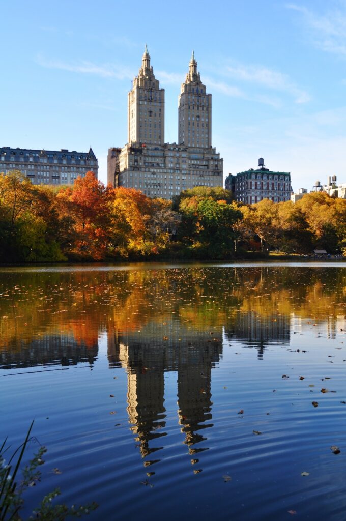 Central Park in NYC in the fall with colorful leaves on the trees and buildings reflected in the pond