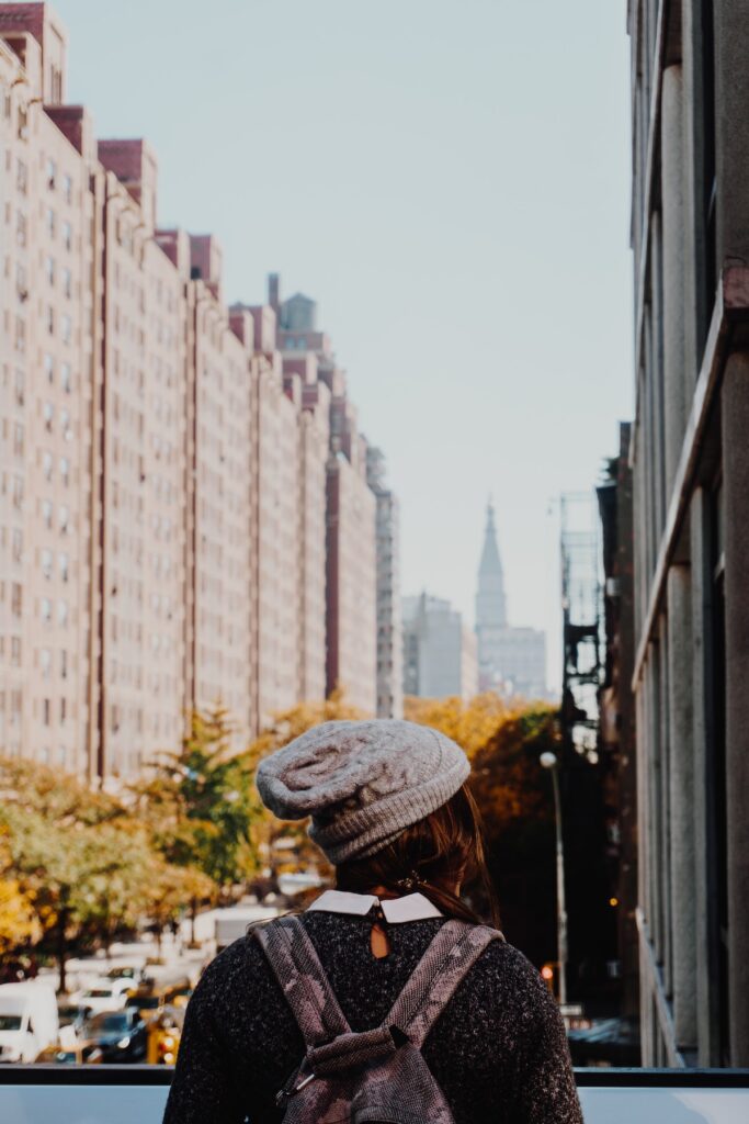 A tourist in NYC in the fall looking over the Highline at the view of buildings and colorful trees, wearing a hat, sweater and backpack