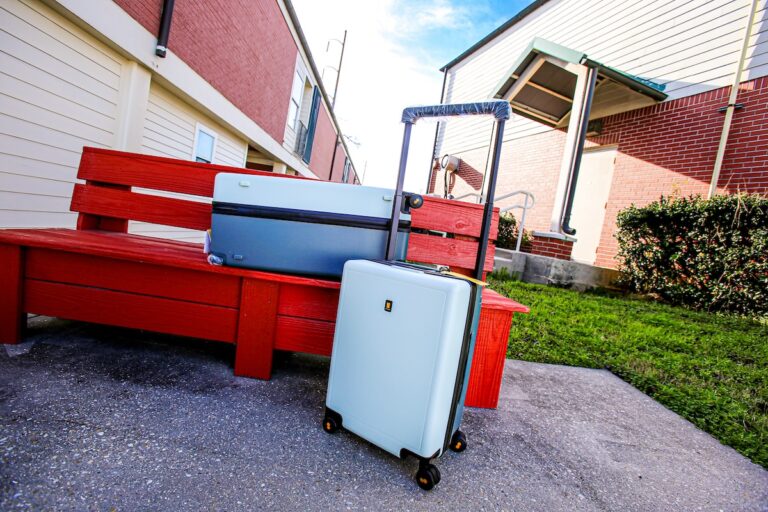 Level8 Voyageur Luggage Set in sage outside on and next to a red painted bench with brick buildings in the background, grass and sky